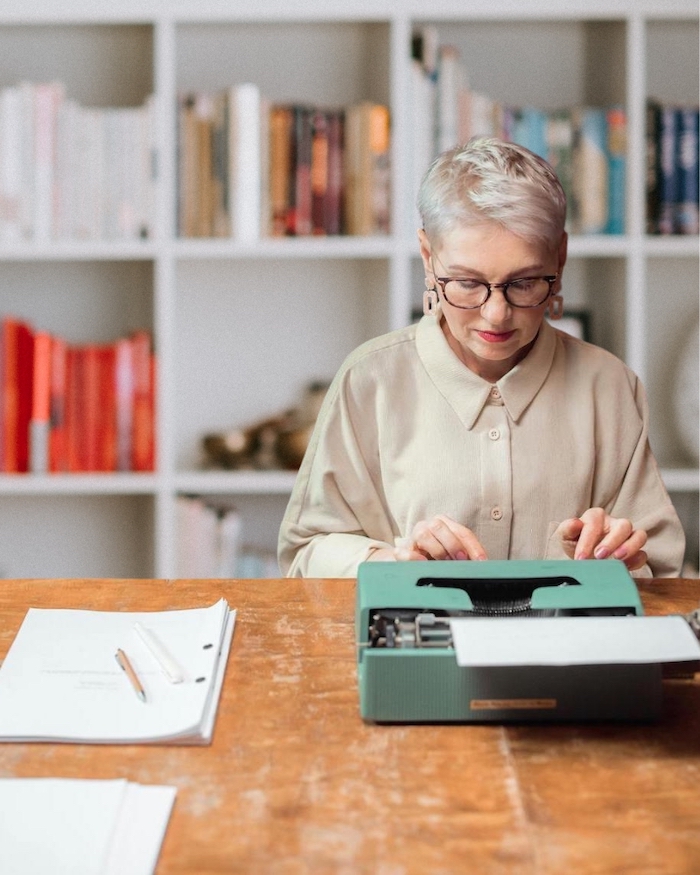 Older woman writing a life story on a green typewriter in front of bookcases.